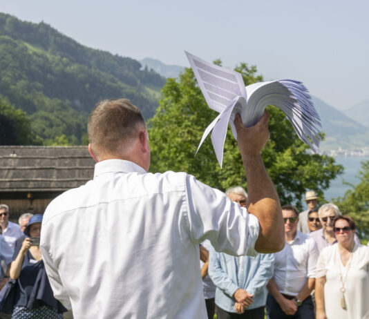 14 juin : le programme extrême de l’UDC pourrait devenir réalité Rassemblement de l'UDC sur la prairie du Grutli : Un homme en chemise blanche tient un modèle de planeur blanc levé vers le ciel, tandis qu'il se tient devant un groupe de personnes près d'un lac de montagne. En arrière-plan, on voit des montagnes boisées, des chalets traditionnels et un lac scintillant.