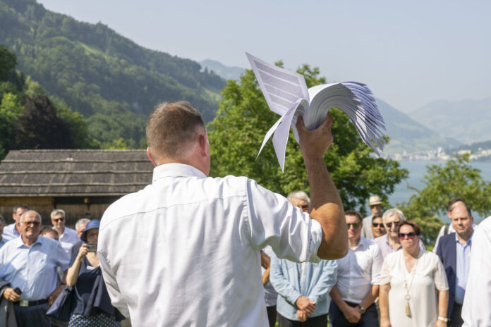 Rassemblement de l'UDC sur la prairie du Grutli : Un homme en chemise blanche tient un modèle de planeur blanc levé vers le ciel, tandis qu'il se tient devant un groupe de personnes près d'un lac de montagne. En arrière-plan, on voit des montagnes boisées, des chalets traditionnels et un lac scintillant.