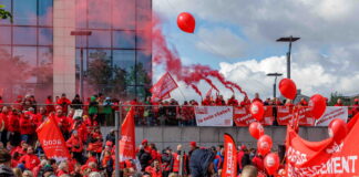 Bonnes nouvelles en mai : la grève reconnue comme droit fondamental, la Belgique résiste à l’austérité et l’ONU adopte une résolution pour le climat Photo montrant une large foule de manifestants lors d'une action syndicale. Les participants, majoritairement vêtus de vestes rouges, brandissent des drapeaux et banderoles aux couleurs des syndicats CGSP, FGTB et CSC. Des ballons rouges flottent dans les airs tandis que des fumigènes rouges sont allumés en arrière-plan. On peut lire sur les banderoles des slogans tels que « Je suis statut », « Touche pas à ma fonction » et « Services publics de qualité », indiquant une mobilisation en défense du secteur public.