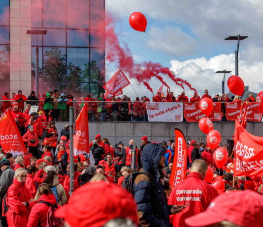 Bonnes nouvelles en mai : la grève reconnue comme droit fondamental, la Belgique résiste à l’austérité et l’ONU adopte une résolution pour le climat Photo montrant une large foule de manifestants lors d'une action syndicale. Les participants, majoritairement vêtus de vestes rouges, brandissent des drapeaux et banderoles aux couleurs des syndicats CGSP, FGTB et CSC. Des ballons rouges flottent dans les airs tandis que des fumigènes rouges sont allumés en arrière-plan. On peut lire sur les banderoles des slogans tels que « Je suis statut », « Touche pas à ma fonction » et « Services publics de qualité », indiquant une mobilisation en défense du secteur public.