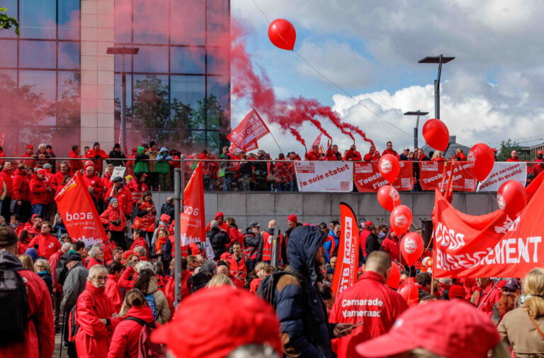 Photo montrant une large foule de manifestants lors d'une action syndicale. Les participants, majoritairement vêtus de vestes rouges, brandissent des drapeaux et banderoles aux couleurs des syndicats CGSP, FGTB et CSC. Des ballons rouges flottent dans les airs tandis que des fumigènes rouges sont allumés en arrière-plan. On peut lire sur les banderoles des slogans tels que « Je suis statut », « Touche pas à ma fonction » et « Services publics de qualité », indiquant une mobilisation en défense du secteur public.