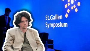 Farbfotografie einer Präsentationsszene beim St.Gallen Symposium. Im Vordergrund sitzt ein etwa vierzigjähriger Mann mit braunem Haar, Schulterlänge, Brille, beige heller Jacke und graugrüner Krawatte, frontal zur Kamera. Im unscharfen Hintergrund steht ein Mann im blauen Anzug. Der Hintergrund ist leuchtendes Blau mit weißem Text „St.Gallen Symposium" und gelben geometrischen Grafikelementen, typisch für die visuelle Identität dieses internationalen Events.