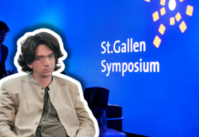 Curtis Yarvin, figure de proue de la droite autoritaire à Saint-Gall Photographie couleur d'une scène de présentation au St.Gallen Symposium. Au premier plan, un homme d'environ quarante ans, cheveux bruns mi-longs, lunettes, vêtu d'une veste beige clair et d'une cravate gris-vert, est assis face à la caméra. À l'arrière-plan flou, un homme en costume bleu se tient debout. Le fond est bleu vif avec le texte blanc « St.Gallen Symposium » et des éléments graphiques géométriques jaunes, typiques de l'identité visuelle de cet événement international.