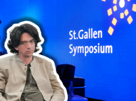 Curtis Yarvin, figure de proue de la droite autoritaire à Saint-Gall Photographie couleur d'une scène de présentation au St.Gallen Symposium. Au premier plan, un homme d'environ quarante ans, cheveux bruns mi-longs, lunettes, vêtu d'une veste beige clair et d'une cravate gris-vert, est assis face à la caméra. À l'arrière-plan flou, un homme en costume bleu se tient debout. Le fond est bleu vif avec le texte blanc « St.Gallen Symposium » et des éléments graphiques géométriques jaunes, typiques de l'identité visuelle de cet événement international.