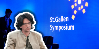 Curtis Yarvin, figure de proue de la droite autoritaire à Saint-Gall Photographie couleur d'une scène de présentation au St.Gallen Symposium. Au premier plan, un homme d'environ quarante ans, cheveux bruns mi-longs, lunettes, vêtu d'une veste beige clair et d'une cravate gris-vert, est assis face à la caméra. À l'arrière-plan flou, un homme en costume bleu se tient debout. Le fond est bleu vif avec le texte blanc « St.Gallen Symposium » et des éléments graphiques géométriques jaunes, typiques de l'identité visuelle de cet événement international.