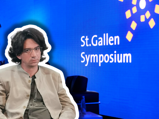 Curtis Yarvin, figure de proue de la droite autoritaire à Saint-Gall Photographie couleur d'une scène de présentation au St.Gallen Symposium. Au premier plan, un homme d'environ quarante ans, cheveux bruns mi-longs, lunettes, vêtu d'une veste beige clair et d'une cravate gris-vert, est assis face à la caméra. À l'arrière-plan flou, un homme en costume bleu se tient debout. Le fond est bleu vif avec le texte blanc « St.Gallen Symposium » et des éléments graphiques géométriques jaunes, typiques de l'identité visuelle de cet événement international.