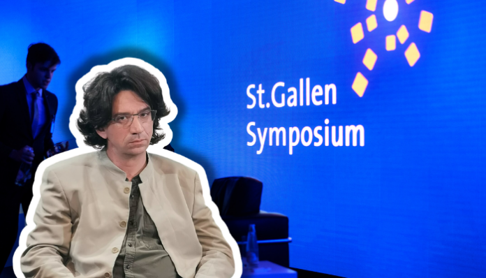 Photographie couleur d'une scène de présentation au St.Gallen Symposium. Au premier plan, un homme d'environ quarante ans, cheveux bruns mi-longs, lunettes, vêtu d'une veste beige clair et d'une cravate gris-vert, est assis face à la caméra. À l'arrière-plan flou, un homme en costume bleu se tient debout. Le fond est bleu vif avec le texte blanc « St.Gallen Symposium » et des éléments graphiques géométriques jaunes, typiques de l'identité visuelle de cet événement international.