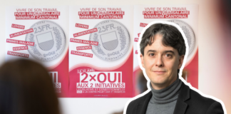 Salaire minimum : « La droite patronale en SManque d’argument » Une personne portant un blazer noir et un col roulé gris se tient devant des affiches de campagne électorale. Les affiches, sur fond rose et blanc, présentent des pièces de monnaie et du texte en français relatif à des initiatives politiques, notamment "2×OUI AUX 2 INITIATIVES". Le message principal porte sur des thèmes sociaux comme le travail, la santé et les allocations familiales, avec mention d'un salaire minimum cantonal.