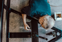 Initiative de l’UDC : « Travailler plus longtemps ? Ce serait réaliste » e plongeante sur un homme âgé aux cheveux gris, portant des lunettes et une chemise bleue, qui manipule des composants électriques ou mécaniques sur une structure en métal noir posée au sol. Des outils et une petite boîte blanche sont visibles à proximité.