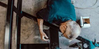 Initiative de l’UDC : « Travailler plus longtemps ? Ce serait réaliste » e plongeante sur un homme âgé aux cheveux gris, portant des lunettes et une chemise bleue, qui manipule des composants électriques ou mécaniques sur une structure en métal noir posée au sol. Des outils et une petite boîte blanche sont visibles à proximité.