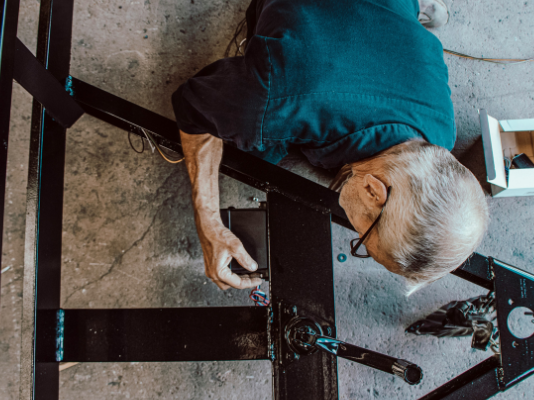 Initiative de l’UDC : « Travailler plus longtemps ? Ce serait réaliste » e plongeante sur un homme âgé aux cheveux gris, portant des lunettes et une chemise bleue, qui manipule des composants électriques ou mécaniques sur une structure en métal noir posée au sol. Des outils et une petite boîte blanche sont visibles à proximité.