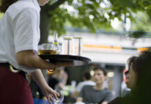 Salaires minimaux : même Guy Parmelin juge la nouvelle loi mauvaise Une serveuse tient un plateau chargé de verres et d'une bouteille Schweppes dans un restaurant en terrasse, avec des clients en arrière-plan.