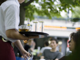 Salaires minimaux : même Guy Parmelin juge la nouvelle loi mauvaise Une serveuse tient un plateau chargé de verres et d'une bouteille Schweppes dans un restaurant en terrasse, avec des clients en arrière-plan.