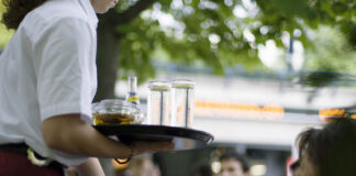 Salaires minimaux : même Guy Parmelin juge la nouvelle loi mauvaise Une serveuse tient un plateau chargé de verres et d'une bouteille Schweppes dans un restaurant en terrasse, avec des clients en arrière-plan.