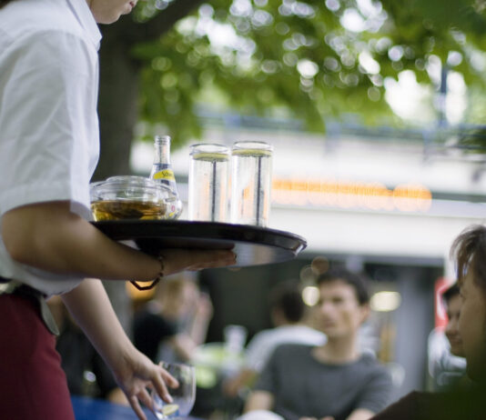 Salaires minimaux : même Guy Parmelin juge la nouvelle loi mauvaise Une serveuse tient un plateau chargé de verres et d'une bouteille Schweppes dans un restaurant en terrasse, avec des clients en arrière-plan.