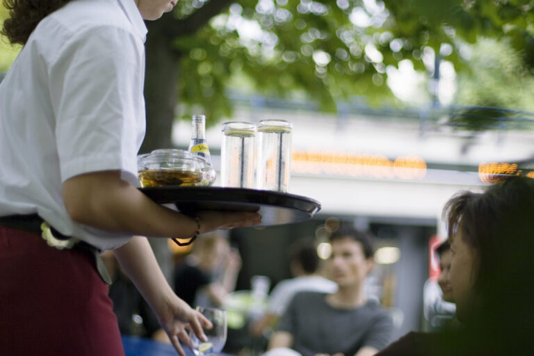 Une serveuse tient un plateau chargé de verres et d'une bouteille Schweppes dans un restaurant en terrasse, avec des clients en arrière-plan.