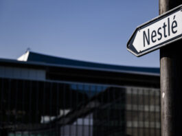 Rapport d’Oxfam sur les multinationales : les actionnaires reçoivent 70 % des bénéfices Panneau de rue indiquant 'Nestlé' en noir et blanc, devant le siège de l'entreprise à Vevey. Un drapeau suisse flotte en arrière-plan sur un bâtiment moderne, sous un ciel bleu.