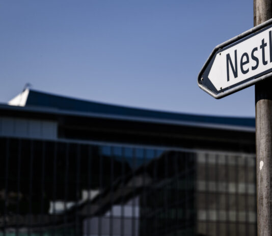 Rapport d’Oxfam sur les multinationales : les actionnaires reçoivent 70 % des bénéfices Panneau de rue indiquant 'Nestlé' en noir et blanc, devant le siège de l'entreprise à Vevey. Un drapeau suisse flotte en arrière-plan sur un bâtiment moderne, sous un ciel bleu.