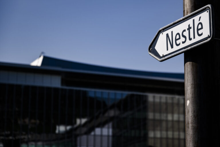 Panneau de rue indiquant 'Nestlé' en noir et blanc, devant le siège de l'entreprise à Vevey. Un drapeau suisse flotte en arrière-plan sur un bâtiment moderne, sous un ciel bleu.