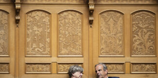 Voici les parlementaires du Centre qui rejettent le gel du salaire minimum Deux personnes en tenue formelle sont assises côte à côte dans un cadre officiel. Elles sont positionnées devant un mur orné de boiseries dorées richement décorées, caractérisé par des panneaux sculptés présentant des motifs floraux et figuratifs élaborés. Les chaises sur lesquelles elles sont assises sont de style classique avec dossiers en rotin. La femme à gauche, vêtue d'une veste noire, regarde vers la droite, tandis que l'homme à droite, portant un costume bleu marine avec une cravate, se tourne légèrement vers elle. L'arrière-plan somptueux suggère un lieu prestigieux tel qu'une institution gouvernementale, un tribunal ou une organisation importante.