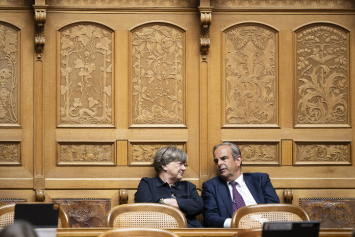 Deux personnes en tenue formelle sont assises côte à côte dans un cadre officiel. Elles sont positionnées devant un mur orné de boiseries dorées richement décorées, caractérisé par des panneaux sculptés présentant des motifs floraux et figuratifs élaborés. Les chaises sur lesquelles elles sont assises sont de style classique avec dossiers en rotin. La femme à gauche, vêtue d'une veste noire, regarde vers la droite, tandis que l'homme à droite, portant un costume bleu marine avec une cravate, se tourne légèrement vers elle. L'arrière-plan somptueux suggère un lieu prestigieux tel qu'une institution gouvernementale, un tribunal ou une organisation importante.