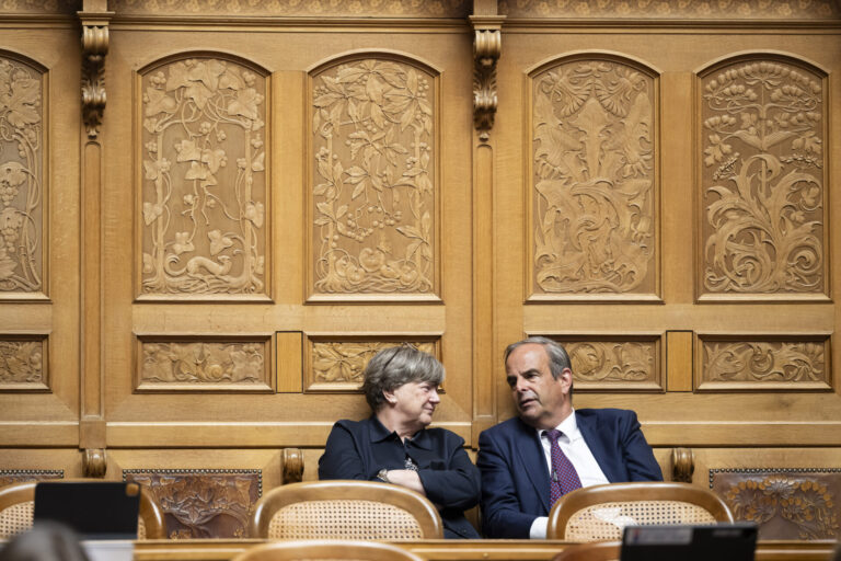 Deux personnes en tenue formelle sont assises côte à côte dans un cadre officiel. Elles sont positionnées devant un mur orné de boiseries dorées richement décorées, caractérisé par des panneaux sculptés présentant des motifs floraux et figuratifs élaborés. Les chaises sur lesquelles elles sont assises sont de style classique avec dossiers en rotin. La femme à gauche, vêtue d'une veste noire, regarde vers la droite, tandis que l'homme à droite, portant un costume bleu marine avec une cravate, se tourne légèrement vers elle. L'arrière-plan somptueux suggère un lieu prestigieux tel qu'une institution gouvernementale, un tribunal ou une organisation importante.