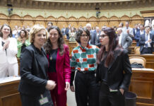 Bonnes nouvelles en juin : résultats de votations réjouissants et succès pour les femmes Quatre femmes politiques se tiennent ensemble dans l'hémicycle d'un parlement, souriantes et engagées dans une conversation. Elles portent des tenues professionnelles variées : l'une en blazer noir, une autre en ensemble rouge fuchsia, la troisième dans un chemisier aux motifs géométriques colorés (vert, orange et blanc), et la dernière en blazer noir. En arrière-plan, l'hémicycle circulaire est rempli de nombreux politiciens et politiciennes en costume-cravate ou tenues formelles. L'architecture classique et élégante du parlement, avec ses arcs dorés et ses boiseries en bois clair, témoigne du caractère solennel du lieu. Cette image illustre la présence croissante des femmes dans les assemblées législatives et leur engagement dans le débat politique.