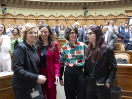 Bonnes nouvelles en juin : résultats de votations réjouissants et succès pour les femmes Quatre femmes politiques se tiennent ensemble dans l'hémicycle d'un parlement, souriantes et engagées dans une conversation. Elles portent des tenues professionnelles variées : l'une en blazer noir, une autre en ensemble rouge fuchsia, la troisième dans un chemisier aux motifs géométriques colorés (vert, orange et blanc), et la dernière en blazer noir. En arrière-plan, l'hémicycle circulaire est rempli de nombreux politiciens et politiciennes en costume-cravate ou tenues formelles. L'architecture classique et élégante du parlement, avec ses arcs dorés et ses boiseries en bois clair, témoigne du caractère solennel du lieu. Cette image illustre la présence croissante des femmes dans les assemblées législatives et leur engagement dans le débat politique.