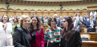 Bonnes nouvelles en juin : résultats de votations réjouissants et succès pour les femmes Quatre femmes politiques se tiennent ensemble dans l'hémicycle d'un parlement, souriantes et engagées dans une conversation. Elles portent des tenues professionnelles variées : l'une en blazer noir, une autre en ensemble rouge fuchsia, la troisième dans un chemisier aux motifs géométriques colorés (vert, orange et blanc), et la dernière en blazer noir. En arrière-plan, l'hémicycle circulaire est rempli de nombreux politiciens et politiciennes en costume-cravate ou tenues formelles. L'architecture classique et élégante du parlement, avec ses arcs dorés et ses boiseries en bois clair, témoigne du caractère solennel du lieu. Cette image illustre la présence croissante des femmes dans les assemblées législatives et leur engagement dans le débat politique.