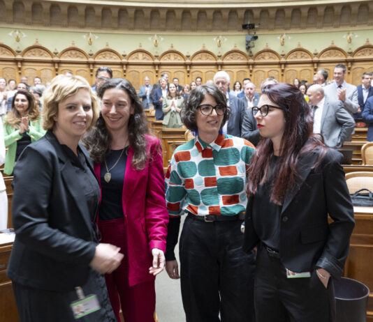 Bonnes nouvelles en juin : résultats de votations réjouissants et succès pour les femmes Quatre femmes politiques se tiennent ensemble dans l'hémicycle d'un parlement, souriantes et engagées dans une conversation. Elles portent des tenues professionnelles variées : l'une en blazer noir, une autre en ensemble rouge fuchsia, la troisième dans un chemisier aux motifs géométriques colorés (vert, orange et blanc), et la dernière en blazer noir. En arrière-plan, l'hémicycle circulaire est rempli de nombreux politiciens et politiciennes en costume-cravate ou tenues formelles. L'architecture classique et élégante du parlement, avec ses arcs dorés et ses boiseries en bois clair, témoigne du caractère solennel du lieu. Cette image illustre la présence croissante des femmes dans les assemblées législatives et leur engagement dans le débat politique.