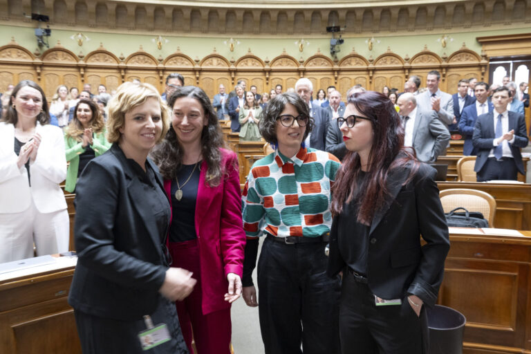 Quatre femmes politiques se tiennent ensemble dans l'hémicycle d'un parlement, souriantes et engagées dans une conversation. Elles portent des tenues professionnelles variées : l'une en blazer noir, une autre en ensemble rouge fuchsia, la troisième dans un chemisier aux motifs géométriques colorés (vert, orange et blanc), et la dernière en blazer noir. En arrière-plan, l'hémicycle circulaire est rempli de nombreux politiciens et politiciennes en costume-cravate ou tenues formelles. L'architecture classique et élégante du parlement, avec ses arcs dorés et ses boiseries en bois clair, témoigne du caractère solennel du lieu. Cette image illustre la présence croissante des femmes dans les assemblées législatives et leur engagement dans le débat politique.