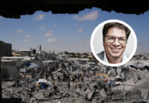« La situation des Palestinien-nes est absolument désespérée » Photographie montrant une personne souriante en portrait inséré sur une vue aérienne d'un camp de réfugiés ou d'une zone densément peuplée et précaires. Le portrait en médaillon, en couleur, représente un homme d'âge moyen aux cheveux courts et grisonnants, portant des lunettes et un veston, qui regarde l'objectif avec un sourire bienveillant. En arrière-plan, en noir et blanc, s'étend un vaste bidonville ou campement informel composé de centaines d'abris de fortune en tôle, bâches et matériaux de récupération. Des allées étroites serpentent entre les constructions, et de nombreuses personnes sont visibles, vaquant à leurs occupations quotidiennes. Le paysage environnant est aride, avec quelques bâtiments en dur dispersés et des collines en arrière-plan sous un ciel bleu clair.