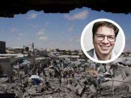 « La situation des Palestinien-nes est absolument désespérée » Photographie montrant une personne souriante en portrait inséré sur une vue aérienne d'un camp de réfugiés ou d'une zone densément peuplée et précaires. Le portrait en médaillon, en couleur, représente un homme d'âge moyen aux cheveux courts et grisonnants, portant des lunettes et un veston, qui regarde l'objectif avec un sourire bienveillant. En arrière-plan, en noir et blanc, s'étend un vaste bidonville ou campement informel composé de centaines d'abris de fortune en tôle, bâches et matériaux de récupération. Des allées étroites serpentent entre les constructions, et de nombreuses personnes sont visibles, vaquant à leurs occupations quotidiennes. Le paysage environnant est aride, avec quelques bâtiments en dur dispersés et des collines en arrière-plan sous un ciel bleu clair.