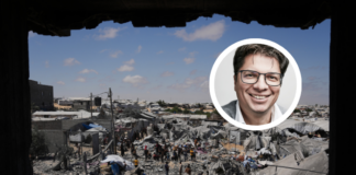 « La situation des Palestinien-nes est absolument désespérée » Photographie montrant une personne souriante en portrait inséré sur une vue aérienne d'un camp de réfugiés ou d'une zone densément peuplée et précaires. Le portrait en médaillon, en couleur, représente un homme d'âge moyen aux cheveux courts et grisonnants, portant des lunettes et un veston, qui regarde l'objectif avec un sourire bienveillant. En arrière-plan, en noir et blanc, s'étend un vaste bidonville ou campement informel composé de centaines d'abris de fortune en tôle, bâches et matériaux de récupération. Des allées étroites serpentent entre les constructions, et de nombreuses personnes sont visibles, vaquant à leurs occupations quotidiennes. Le paysage environnant est aride, avec quelques bâtiments en dur dispersés et des collines en arrière-plan sous un ciel bleu clair.