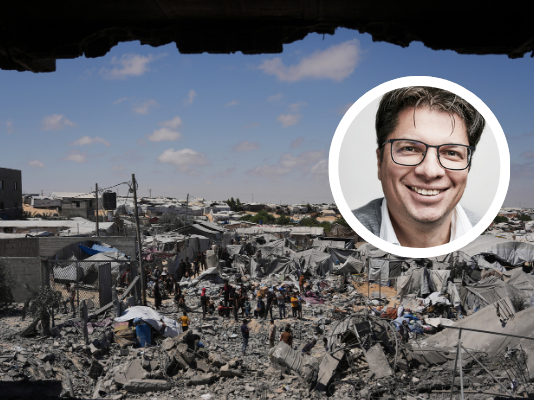 « La situation des Palestinien-nes est absolument désespérée » Photographie montrant une personne souriante en portrait inséré sur une vue aérienne d'un camp de réfugiés ou d'une zone densément peuplée et précaires. Le portrait en médaillon, en couleur, représente un homme d'âge moyen aux cheveux courts et grisonnants, portant des lunettes et un veston, qui regarde l'objectif avec un sourire bienveillant. En arrière-plan, en noir et blanc, s'étend un vaste bidonville ou campement informel composé de centaines d'abris de fortune en tôle, bâches et matériaux de récupération. Des allées étroites serpentent entre les constructions, et de nombreuses personnes sont visibles, vaquant à leurs occupations quotidiennes. Le paysage environnant est aride, avec quelques bâtiments en dur dispersés et des collines en arrière-plan sous un ciel bleu clair.
