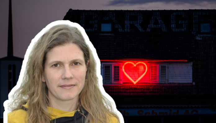Une femme aux cheveux longs, châtain clair et ondulés, portant une veste sombre et un haut jaune, est représentée en premier plan. En arrière-plan, on distingue un bâtiment à toit sombre sur lequel une enseigne lumineuse rouge en forme de cœur est projetée ou affichée sur une fenêtre à volets. L'atmosphère générale de l'image est sombre, contrastant avec la lumière rouge vive du cœur.