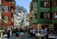 Démantèlement du droit du travail : une commission souhaite étendre massivement les horaires d’ouverture le dimanche Rue piétonne à Zurich, colorée d'une vieille ville suisse, probablement Lucerne. Des bâtiments historiques aux façades peintes dans des teintes vives - rouge, orange, vert, jaune et bleu - bordent la rue pavée. Sur la droite, un établissement nommé "Cantinetta Antinori" accueille des clients assis à des tables extérieures. De nombreux piétons flânent dans cette atmosphère touristique ensoleillée, sous un ciel bleu clair. Les fenêtres et les volets colorés des immeubles anciens témoignent du charme architectural typique des centres-villes suisses médiévaux.