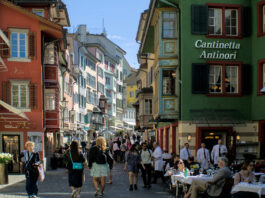 Démantèlement du droit du travail : une commission souhaite étendre massivement les horaires d’ouverture le dimanche Rue piétonne à Zurich, colorée d'une vieille ville suisse, probablement Lucerne. Des bâtiments historiques aux façades peintes dans des teintes vives - rouge, orange, vert, jaune et bleu - bordent la rue pavée. Sur la droite, un établissement nommé "Cantinetta Antinori" accueille des clients assis à des tables extérieures. De nombreux piétons flânent dans cette atmosphère touristique ensoleillée, sous un ciel bleu clair. Les fenêtres et les volets colorés des immeubles anciens témoignent du charme architectural typique des centres-villes suisses médiévaux.