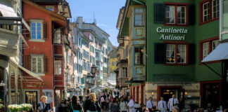 Démantèlement du droit du travail : une commission souhaite étendre massivement les horaires d’ouverture le dimanche Rue piétonne à Zurich, colorée d'une vieille ville suisse, probablement Lucerne. Des bâtiments historiques aux façades peintes dans des teintes vives - rouge, orange, vert, jaune et bleu - bordent la rue pavée. Sur la droite, un établissement nommé "Cantinetta Antinori" accueille des clients assis à des tables extérieures. De nombreux piétons flânent dans cette atmosphère touristique ensoleillée, sous un ciel bleu clair. Les fenêtres et les volets colorés des immeubles anciens témoignent du charme architectural typique des centres-villes suisses médiévaux.