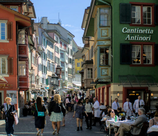 Démantèlement du droit du travail : une commission souhaite étendre massivement les horaires d’ouverture le dimanche Rue piétonne à Zurich, colorée d'une vieille ville suisse, probablement Lucerne. Des bâtiments historiques aux façades peintes dans des teintes vives - rouge, orange, vert, jaune et bleu - bordent la rue pavée. Sur la droite, un établissement nommé "Cantinetta Antinori" accueille des clients assis à des tables extérieures. De nombreux piétons flânent dans cette atmosphère touristique ensoleillée, sous un ciel bleu clair. Les fenêtres et les volets colorés des immeubles anciens témoignent du charme architectural typique des centres-villes suisses médiévaux.