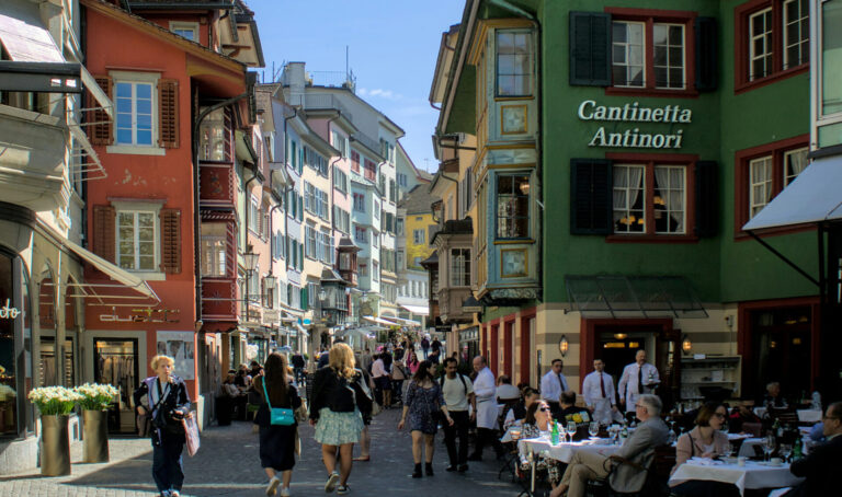 Rue piétonne à Zurich, colorée d'une vieille ville suisse, probablement Lucerne. Des bâtiments historiques aux façades peintes dans des teintes vives - rouge, orange, vert, jaune et bleu - bordent la rue pavée. Sur la droite, un établissement nommé "Cantinetta Antinori" accueille des clients assis à des tables extérieures. De nombreux piétons flânent dans cette atmosphère touristique ensoleillée, sous un ciel bleu clair. Les fenêtres et les volets colorés des immeubles anciens témoignent du charme architectural typique des centres-villes suisses médiévaux.