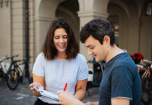 Énergie nucléaire, salaires minimaux et Malaisie : ces trois référendums sont prêts à être signés Deux jeunes adultes souriants examinent ensemble un document ou formulaire dans une cour historique aux arcades. La femme en t-shirt clair tient le papier tandis que l'homme en t-shirt bleu foncé y ajoute une signature avec un stylo rouge. Des vélos sont garés en arrière-plan contre les murs en pierre.