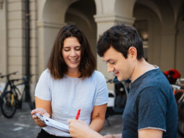 Énergie nucléaire, salaires minimaux et Malaisie : ces trois référendums sont prêts à être signés Deux jeunes adultes souriants examinent ensemble un document ou formulaire dans une cour historique aux arcades. La femme en t-shirt clair tient le papier tandis que l'homme en t-shirt bleu foncé y ajoute une signature avec un stylo rouge. Des vélos sont garés en arrière-plan contre les murs en pierre.
