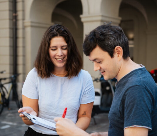 Énergie nucléaire, salaires minimaux et Malaisie : ces trois référendums sont prêts à être signés Deux jeunes adultes souriants examinent ensemble un document ou formulaire dans une cour historique aux arcades. La femme en t-shirt clair tient le papier tandis que l'homme en t-shirt bleu foncé y ajoute une signature avec un stylo rouge. Des vélos sont garés en arrière-plan contre les murs en pierre.