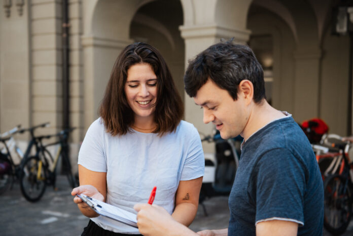 Deux jeunes adultes souriants examinent ensemble un document ou formulaire dans une cour historique aux arcades. La femme en t-shirt clair tient le papier tandis que l'homme en t-shirt bleu foncé y ajoute une signature avec un stylo rouge. Des vélos sont garés en arrière-plan contre les murs en pierre.