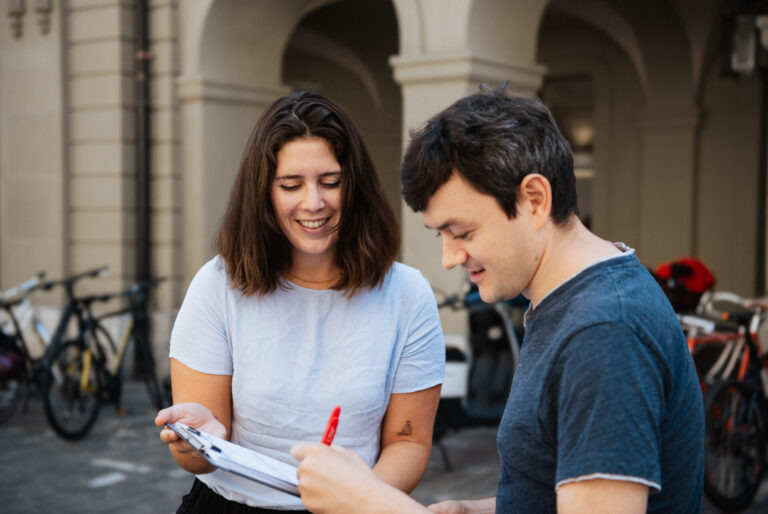 Deux jeunes adultes souriants examinent ensemble un document ou formulaire dans une cour historique aux arcades. La femme en t-shirt clair tient le papier tandis que l'homme en t-shirt bleu foncé y ajoute une signature avec un stylo rouge. Des vélos sont garés en arrière-plan contre les murs en pierre.