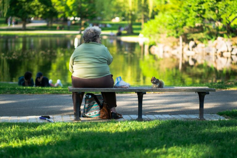 Une femme âgée assise sur un banc dans un parc, nourrissant un écureuil près d'un étang. Arrière-plan verdoyant et ensoleillé.
