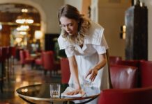 Ce qu’il faut savoir sur l’attaque de la droite contre le salaire minimum Une serveuse en uniforme blanc dessert une table dans un élégant salon d'hôtel ou de restaurant. Elle place un verre d'eau sur une table ronde noire, avec professionnalisme et attention. L'arrière-plan montre un intérieur raffiné avec des fauteuils rouges, un éclairage tamisé et une architecture classique. L'ambiance générale reflète un service hôtelier de qualité dans un établissement luxueux.
