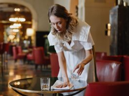 Ce qu’il faut savoir sur l’attaque de la droite contre le salaire minimum Une serveuse en uniforme blanc dessert une table dans un élégant salon d'hôtel ou de restaurant. Elle place un verre d'eau sur une table ronde noire, avec professionnalisme et attention. L'arrière-plan montre un intérieur raffiné avec des fauteuils rouges, un éclairage tamisé et une architecture classique. L'ambiance générale reflète un service hôtelier de qualité dans un établissement luxueux.