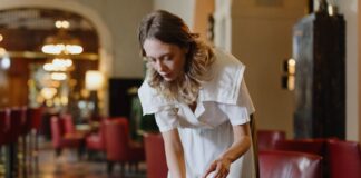 Ce qu’il faut savoir sur l’attaque de la droite contre le salaire minimum Une serveuse en uniforme blanc dessert une table dans un élégant salon d'hôtel ou de restaurant. Elle place un verre d'eau sur une table ronde noire, avec professionnalisme et attention. L'arrière-plan montre un intérieur raffiné avec des fauteuils rouges, un éclairage tamisé et une architecture classique. L'ambiance générale reflète un service hôtelier de qualité dans un établissement luxueux.